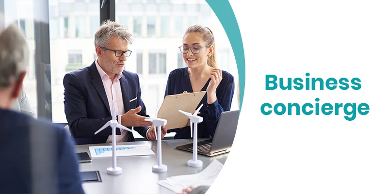 Two people in an office setting, one holding a clipboard and smiling, discuss business concierge services over a table adorned with wind turbine models. A laptop and documents are visible. The text Business concierge is displayed on the right side of the image.