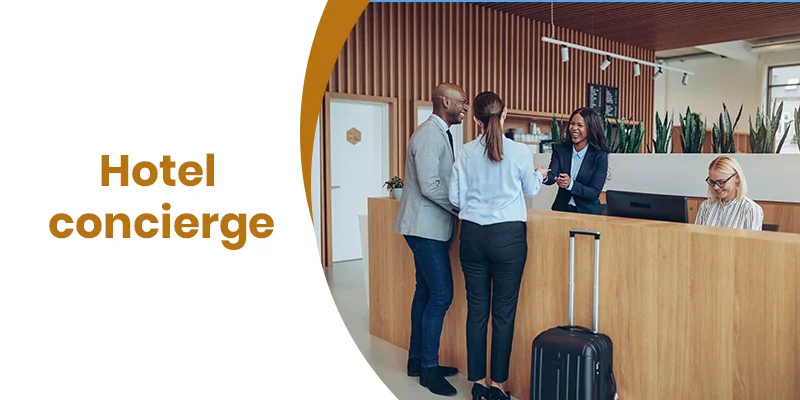 A hotel concierge desk with two guests being assisted by a staff member showcases modern design, with Hotel Concierge Services displayed on the left. A suitcase is nearby while another team member efficiently works at a computer, ensuring seamless guest experiences.